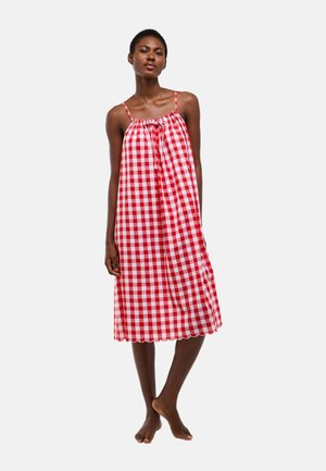 Femme debout pieds nus portant une robe ample à carreaux rouges et blancs sans manches avec de fines bretelles sur un fond uni.