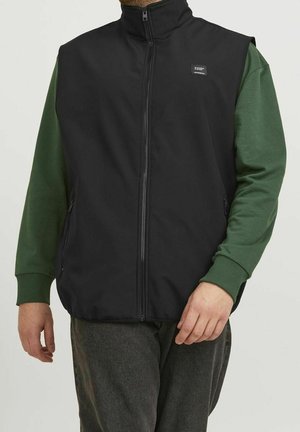 Mężczyzna noszący czarną bezrękawnik na zamek błyskawiczny na zieloną koszulę z długim rękawem i ciemne spodnie, stojący ze swobodnie opartymi ramionami wzdłuż ciała.