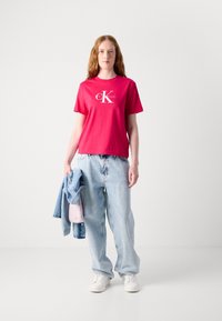 Camiseta de algodón roja con un logo blanco de CK, combinada con unos vaqueros holgados de color azul claro y una chaqueta de denim. El modelo sostiene un bolso rosa.