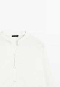 Hvid skjorte med knapper og ståkrave, lavet af glat stof, med en frontplade med knapper og lange ærmer.