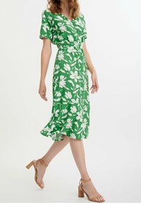 Grön blommig klänning med V-ringning, korta ärmar och elastisk midja. Har vita blommönster och en flödande fåll. Matchas med tanfärgade blockklackssandaletter.