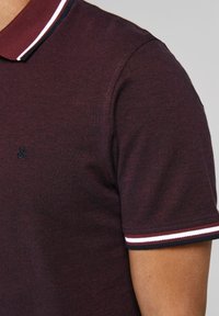 Burgundin värinen polo-paita, jossa on teksturoitu viimeistely, sisältää punaisia ja valkoisia raitoja kauluksessa, ja rinnassa on pieni musta logo.