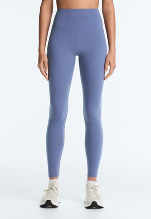 Femme portant des leggings bleus taille haute et des baskets blanches, debout droite devant un fond blanc uni.