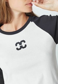 Baltos ir juodos spalvos raglan tipo marškiniai su apvaliu kaklu ir trumpomis rankovėmis; krūtinės srityje yra juodas logotipas. Sklandus medvilnės audinys.