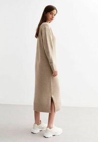 Beige gebreide jurk met lange mouwen, ribbelmanchetten en een zijsplit. Draagt witte sportschoenen op een lichtgrijze vloer.