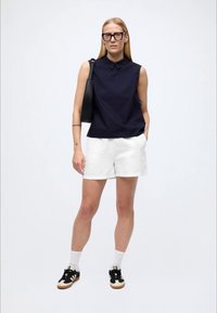 Μπλε σκούρο αμάνικο πουκάμισο με γιακά, λευκά σορτς από λινό, μαύρα αθλητικά παπούτσια με κρεμ λεπτομέρειες και μαύρη τσάντα με μίνιμαλ σχεδιασμό.