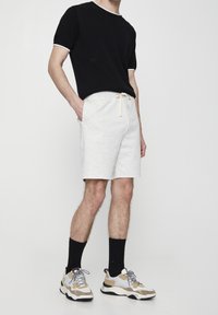 T-shirt noir à col rond et manches courtes avec des détails blancs, accompagné de shorts gris clair à cordon de serrage. Les chaussures présentent un mélange de couleurs beige et blanche.