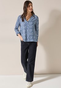 Blå mönstrad blus med V-ringning och långa ärmar, i kombination med mörka vida jeans och vita sneakers.