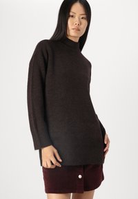 Donkerbruin, oversized gebreide trui met een hoge kraag, geribde manchetten en een zachte textuur, gecombineerd met bordeauxrode corduroy shorts.