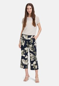 Jasno-beżowa siateczkowa bluzka z krótkim rękawem w połączeniu z granatowymi spodniami 3/4 w kwiaty, z białymi i zielonymi wzorami liści. Modelka nosi przezroczyste sandały na obcasie.