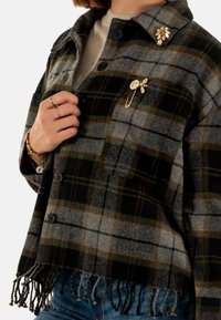 Chaqueta de franela a cuadros en tonos de gris, negro y verde oliva. Presenta un dobladillo con flecos y pines decorativos en el cuello.