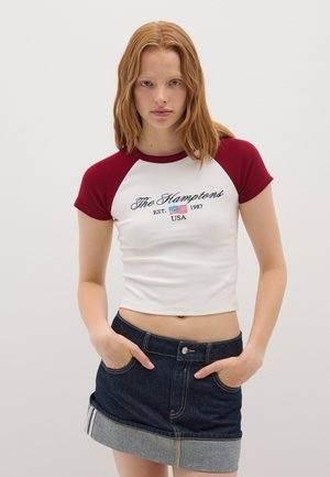 Top crop blanco de canalé con mangas raglán burdeos, que presenta la inscripción "The Hamptons EST. 1987 USA" en bordado azul. Combinado con una falda de mezclilla.