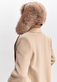 Beige villakangastakki, jossa on pehmeä, pörröinen ruskea turkkihuppu. Varustettu kauluksella ja ribbiohjaimilla kaula-alueella. Takaa otettu kuva näkyy.