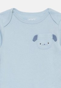 Hellblaues Baumwoll-Overall mit einer kleinen Tasche, die ein gesticktes Hundedesign mit Ohren und Gesichtszügen zeigt. Rundhalsausschnitt.