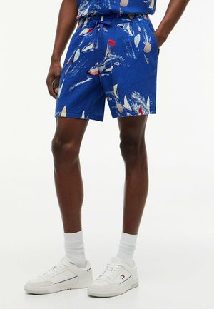 Hombre con pantalones cortos azules con estampados coloridos de veleros, calcetines blancos y zapatillas blancas, de pie con las manos en los bolsillos frente a un fondo blanco.