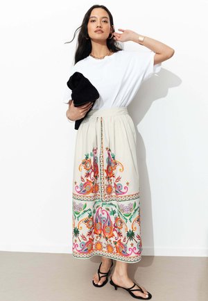 Femme portant un t-shirt blanc, longue jupe crème avec broderie florale colorée, sandales noires, tenant un vêtement noir plié sur fond blanc.