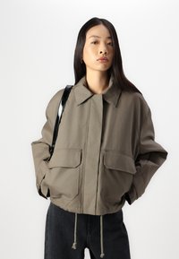 Giacca cropped color khaki con vestibilità ampia, grandi tasche frontali e colletto piatto. Presenta un orlo con coulisse e una tracolla nera lucida.