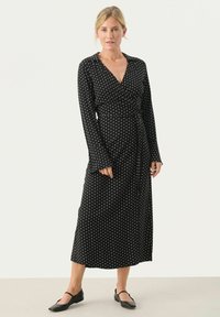 Schwarzes Wickelkleid mit weißen Punkten, langen Ärmeln und gebundenem Taillenbund. Sanfter Stoff mit fließender Silhouette, die bis zur Mitte der Wade reicht. Schwarze flache Schuhe.