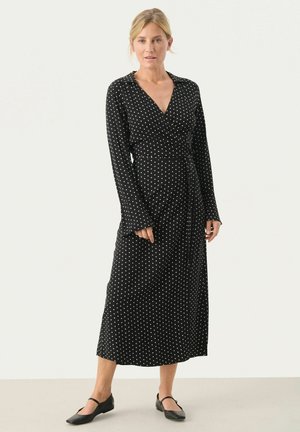Schwarzes Wickelkleid mit weißen Punkten, langen Ärmeln und gebundenem Taillenbund. Sanfter Stoff mit fließender Silhouette, die bis zur Mitte der Wade reicht. Schwarze flache Schuhe.