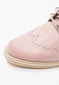 Pointe de chaussure brogue en daim rose avec semelle beige et perforations décoratives, partiellement visible sur fond blanc.