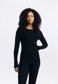 Mulher com cabelo encaracolado, vestindo uma blusa preta de manga longa ajustada e calças pretas, de pé com uma mão na cintura contra um fundo branco simples.