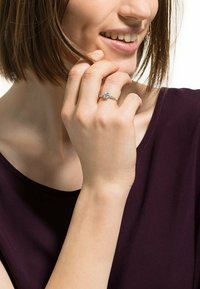 Anello d'oro con una pietra preziosa centrale trasparente, banda ornata e dettagli delicati. Modella che tiene l'anello con una mano rilassata vicino al viso.