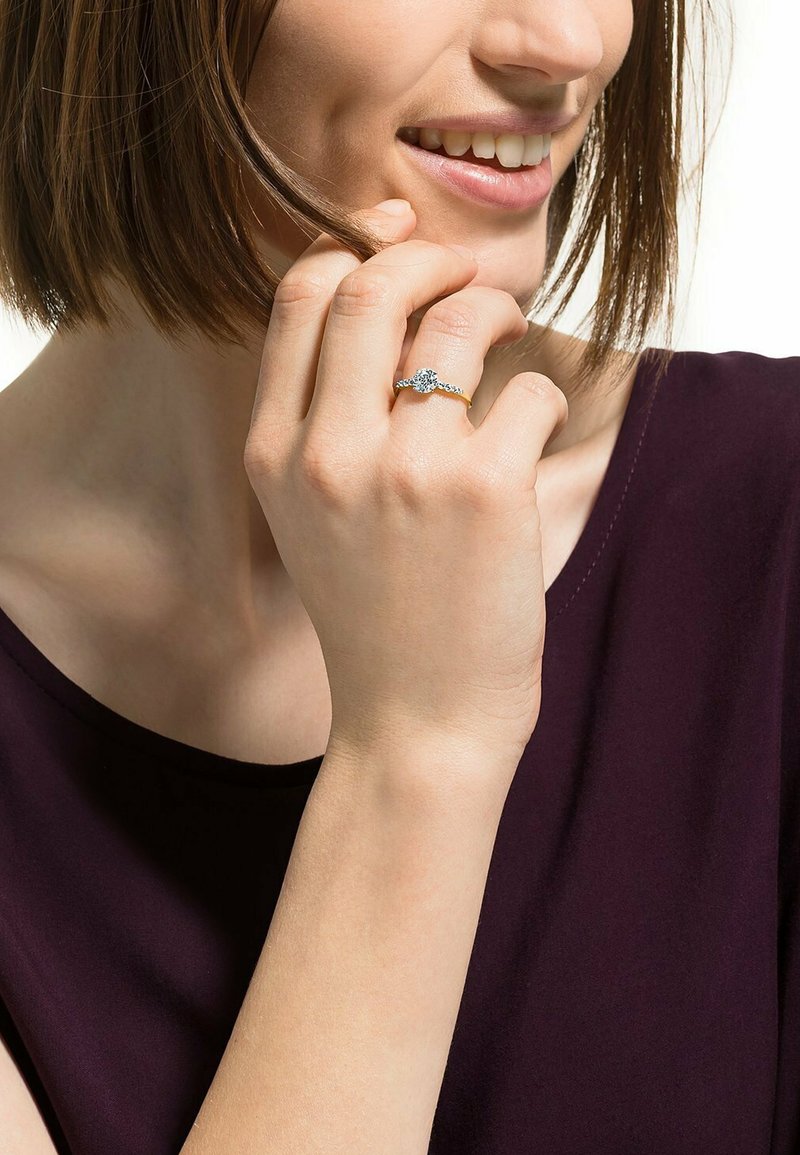 Anello d'oro con una pietra preziosa centrale trasparente, banda ornata e dettagli delicati. Modella che tiene l'anello con una mano rilassata vicino al viso.