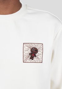 Felpa bianca con un patch ricamato marrone raffigurante un personaggio dei cartoni animati e la scritta "WRONG FRIENDS" su uno sfondo a motivi.