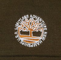 Beanie verde oliva con texture a coste; presenta un logo ricamato circolare in bianco e arancione che raffigura un albero e il testo "AVVENTURA SENZA LIMITI."