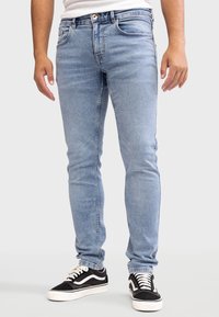 Vaqueros de mezclilla azul claro con un corte ceñido, que cuentan con cierre de botón, cinco bolsillos y un ligero desgaste. Combinados con zapatillas negras y blancas.