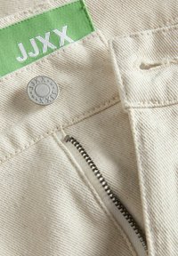 Bežasti pamučni materijal s vidljivim patentnim zatvaračem, metalnim kopčanjem i zelenom oznakom koja prikazuje "JJXX" u bijelom tekstu.