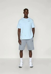 Šviesiai mėlynas medvilninis marškinėliai su Nike logotipu ir spalvinga dėmių dizainu, derinami su pilkomis šortais ir baltais sportiniais batais.