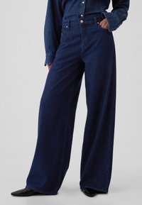 Sötétkék farmert széles szárú nadrág, magas derékkal, elülső zsebekkel és laza szabással. Finoman varrott, klasszikus kialakítással.