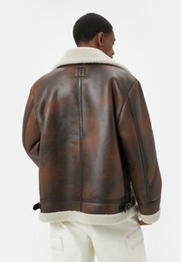 Giacca in shearling marrone con colletto bianco spesso, zip frontale e fibbie regolabili all'orlo. Presenta una finitura testurizzata e una vestibilità rilassata.