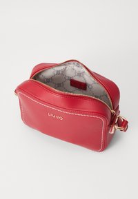 Borsa a tracolla in pelle rossa con hardware dorato e cuciture bianche; chiusura con zip; l'interno presenta una fodera a motivo e dettagli del logo.