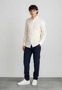 Kremasta košulja na dugmad s čvrstim ovratnikom, uparena s tamnoplavim hlačama i bijelim sportskim cipelama. Minimalna tekstura, prilagođen kroj.