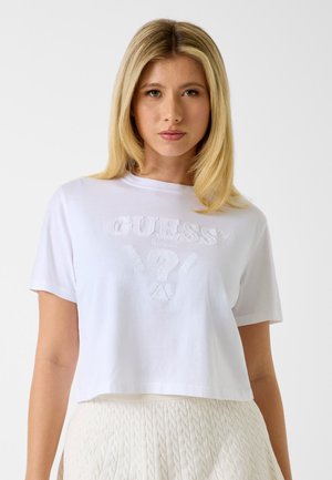 Donna con capelli biondi che indossa una t-shirt bianca corta di Guess con logo in rilievo e una gonna bianca testurizzata, in piedi davanti a uno sfondo semplice.