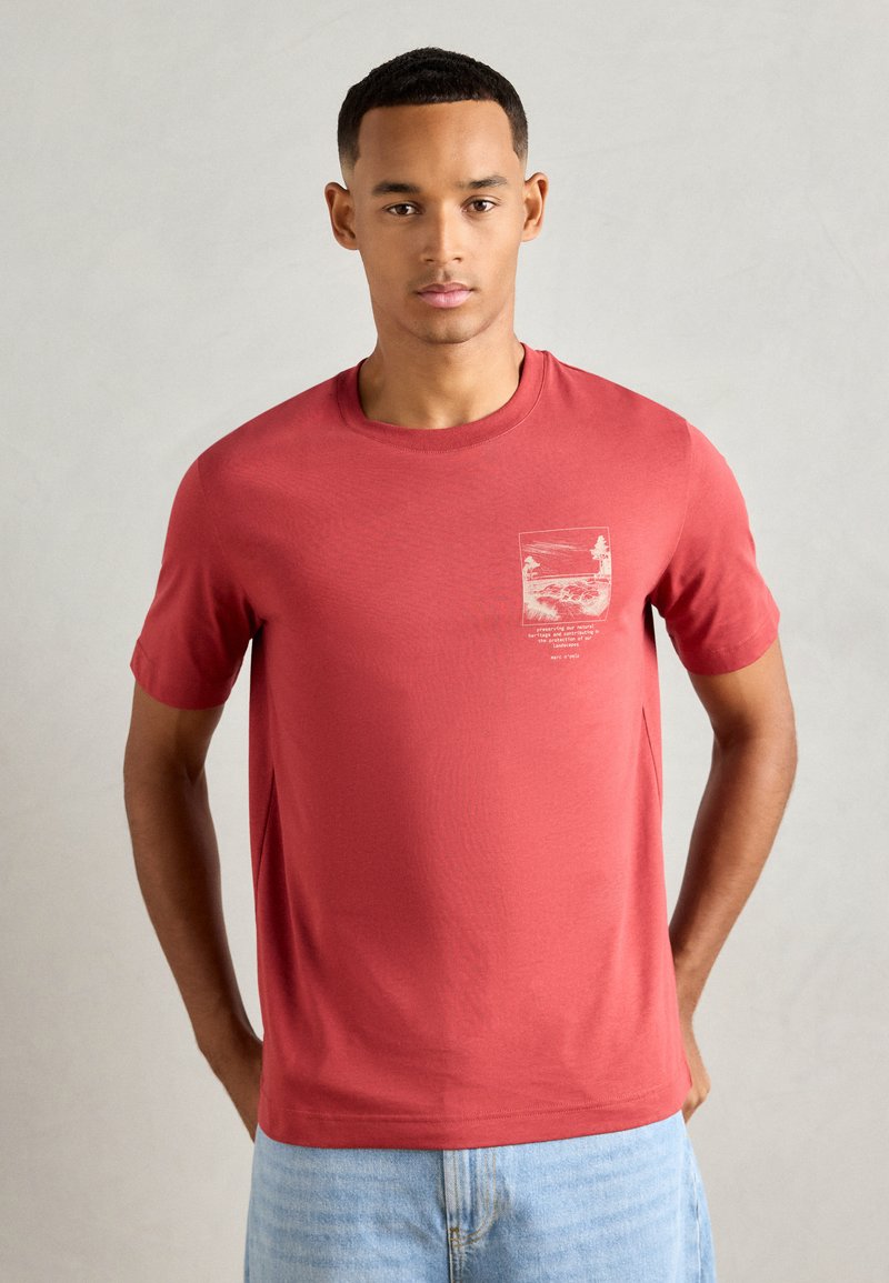 Moški nosi kratkosrajno rdečo majico z lahkim grafičnim oblikovanjem in besedilom na prsih, v kombinaciji z svetlobno modrimi kavbojkami, stoji ob običajni ozadju.