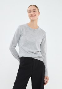 Szary sweter dzianinowy z długimi rękawami i okrągłym dekoltem, w połączeniu z czarnymi spodniami. Gładka struktura z prążkowanymi detalami przy mankietach i dolnym zakończeniu.