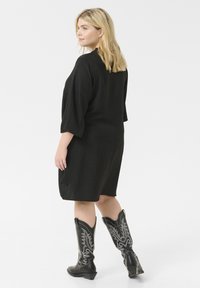 Robe noire mi-longue avec des manches trois-quarts, en tissu texturé, décolleté rond, portée avec des bottes de cowboy noires ornées d'une broderie en argent complexe.