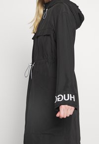 Parka impermeabile nero con cappuccio, vita regolabile con coulisse, tasche laterali e testo bianco con marchio sulla manica. Texture liscia.