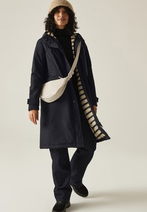 Woman wearing a beige knit hat, navy coat with striped lining, black pants, black shoes, and a beige crossbody bag walking against a plain background.