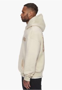 Beige hettegenser laget av myk stoff, med en stor hette, sidelomme og en brun logo-detalj. Avslappet passform med ribbestrikkede mansjetter.