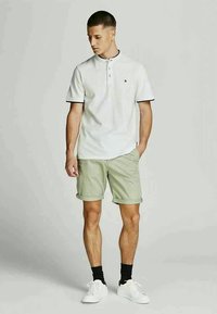 Man wearing white short-sleeve polo shirt, light green rolled-up shorts, black socks, and white sneakers, standing with one hand in pocket.