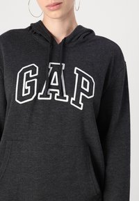 Hanorac gri închis din materiale moi, cu un logo mare brodat "GAP" în alb. Include o buzunar frontal și glugă cu șnur.