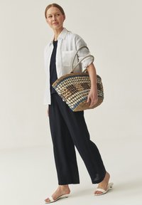 Woven straw bag with blue and white accents, featuring curved handles. Model wears a white shirt and navy pants, with white slide sandals.