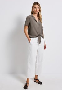 Blusa khaki a maniche corte con dettaglio al centro, abbinata a pantaloni bianchi a gamba larga e sandali neri intrecciati. Tessuto di finitura liscia.