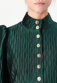 Close-up of a person wearing a dark green textured blouse with a high collar and seven vertical gold buttons.
