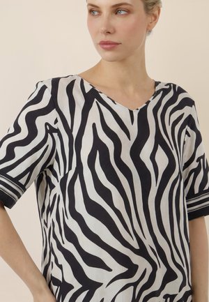 Frau trägt ein schwarz-weiß gestreiftes Zebra-Muster V-Ausschnitt-Oberteil mit kurzen Ärmeln, schaut seitlich vor einem schlichten Hintergrund.