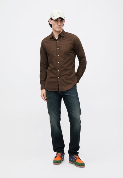 Hombre con gorra beige, camisa marrón de botones, jeans azul oscuro y zapatos multicolores en verde, naranja y azul, de pie frente a un fondo blanco.